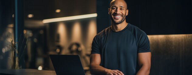 ein männlicher Fitnesstrainer steht hinter einem Steintresen mit einem Laptop in einem Fitnessstudio und lächelt in die Kamera