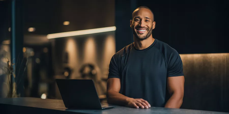 ein männlicher Fitnesstrainer steht hinter einem Steintresen mit einem Laptop in einem Fitnessstudio und lächelt in die Kamera
